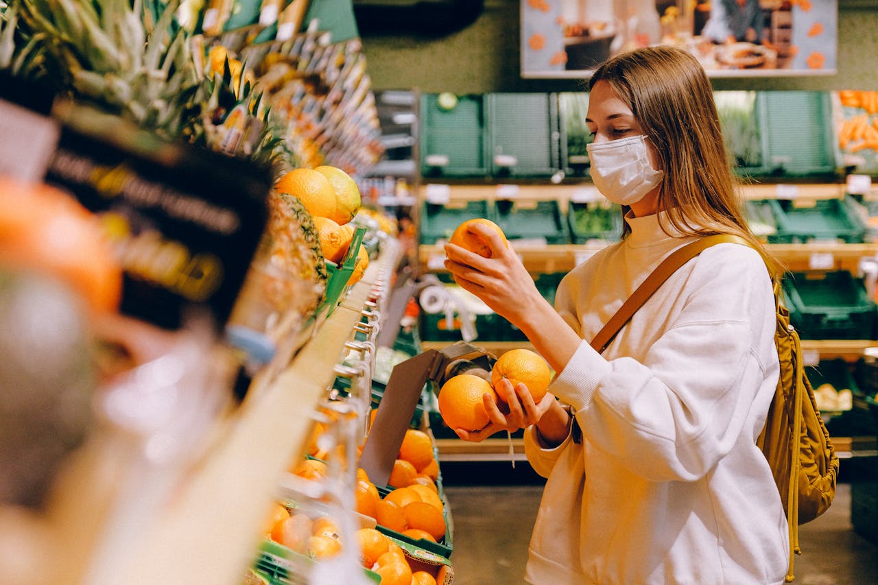 A woman wearing a face mask selects oranges in a grocery store, focusing on safety and prevention.